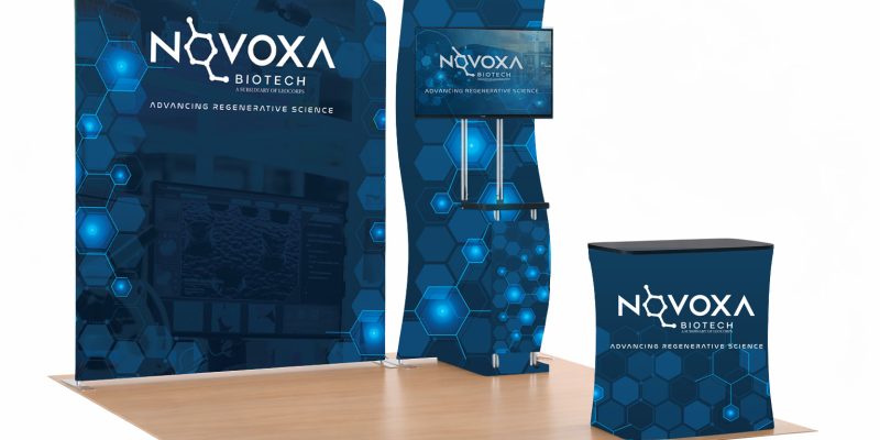 Trade show booth for NovoVoxa Biotech with a blue hex pattern backdrop, tall display, monitor, and matching counter on a wooden floor.